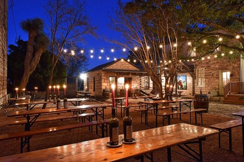 Outdoor dining area at dusk, featuring wooden tables, candles, and string lights, with a rustic building in the background.