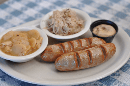 A plate with two grilled sausages, sauerkraut, apple sauce, and a small cup of mustard on a checkered tablecloth.