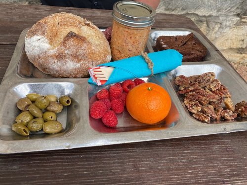 A metal tray with bread, a jar of spread, raspberries, an orange, olives, and candied pecans.