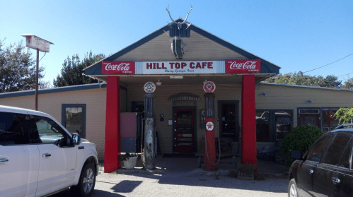 Exterior of Hill Top Cafe featuring a vintage sign, red accents, and classic gas pumps in front.