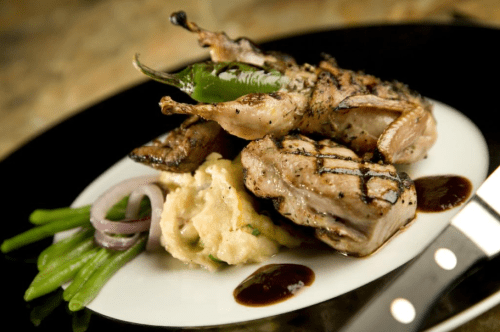 Grilled quail served with mashed potatoes, green beans, and a jalapeño, drizzled with sauce on a white plate.