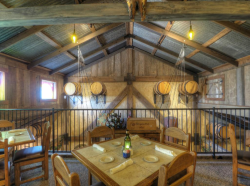 Cozy rustic dining area with wooden tables, chairs, and barrels, featuring warm lighting and a charming atmosphere.