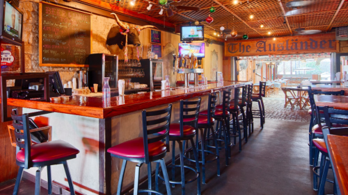 A rustic bar interior with wooden counters, high stools, and festive decorations, featuring a chalkboard menu and TVs.