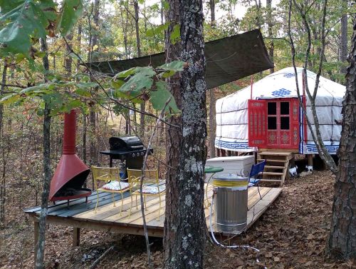 A cozy yurt nestled in the woods, with a wooden deck, outdoor seating, and a grill under a shaded canopy.