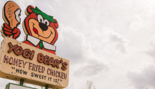 Sign for Yogi Bear's Honey Fried Chicken featuring a cartoon bear holding a drumstick, with a cloudy sky in the background.