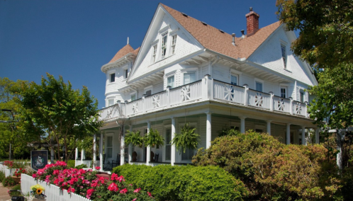 A charming white Victorian house with a wraparound porch, surrounded by vibrant flowers and greenery under a clear blue sky.
