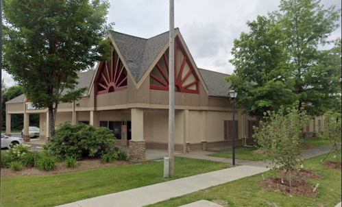 A modern building with a unique roof design, surrounded by trees and landscaping, on a cloudy day.