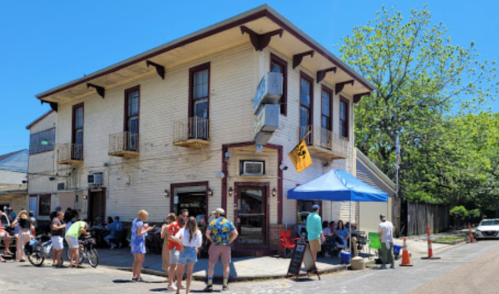 A busy street scene featuring a two-story building with people gathered outside under a blue tent on a sunny day.