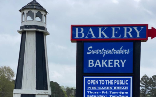 A bakery sign with a lighthouse structure nearby, indicating hours and offerings like pies, cakes, and bread.