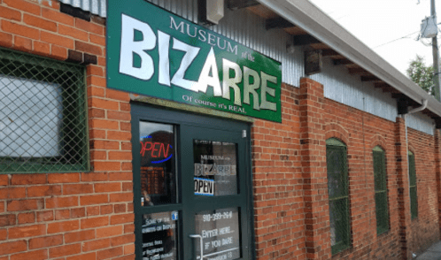 Exterior of the Museum of the Bizarre, featuring a green sign and brick walls, with an "Open" neon sign in the window.