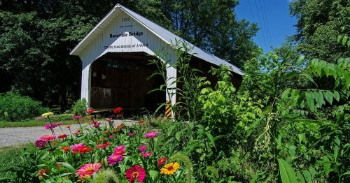 A covered bridge surrounded by vibrant flowers and greenery under a clear blue sky.