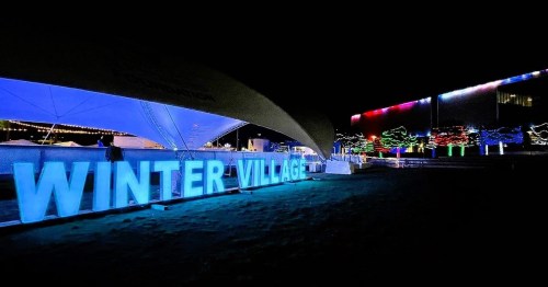 Brightly lit "WINTER VILLAGE" sign at night, surrounded by colorful lights and festive decorations.