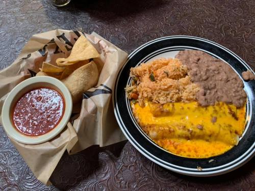 A plate of enchiladas topped with cheese, served with rice, refried beans, and a side of tortilla chips and salsa.