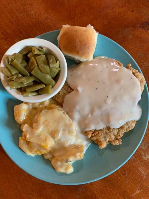 A plate of fried chicken with gravy, macaroni and cheese, green beans, and a roll on a blue plate.