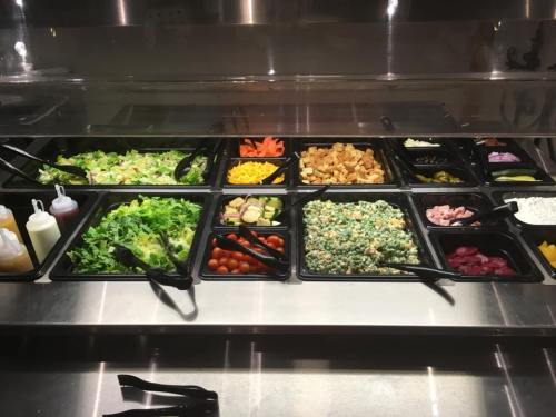 A variety of fresh salads and toppings displayed in black containers on a buffet counter.