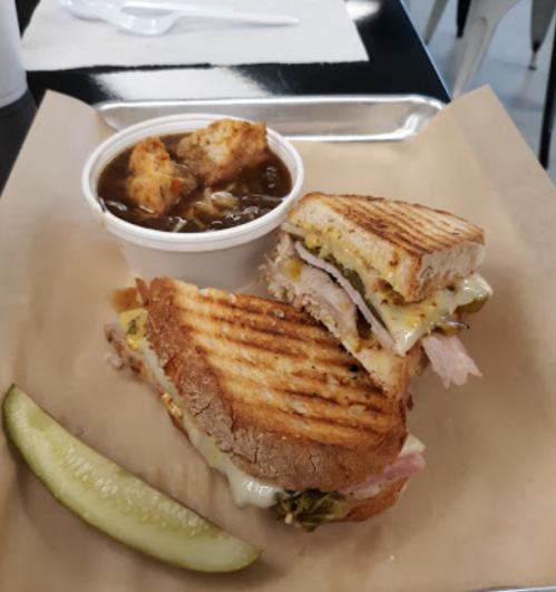 A grilled sandwich with turkey and cheese, served with a cup of soup and a pickle slice on a tray.