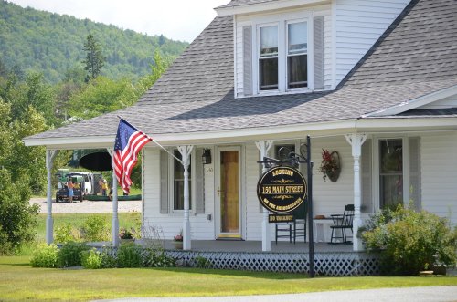 A charming white house with a porch, an American flag, and a sign reading "Lodging 150 Main Street" in a scenic setting.