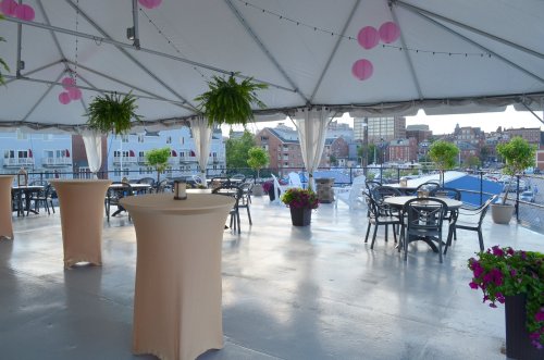 A spacious outdoor event tent with tables, plants, and a view of a marina and city skyline.