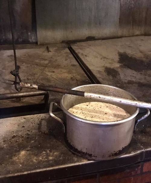 A metal pot with a dark liquid sits on a stove, alongside a long metal stirring rod in a rustic kitchen setting.