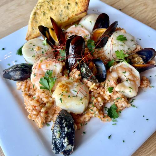 A plate of seafood risotto with shrimp, scallops, mussels, and a slice of garlic bread, garnished with herbs.