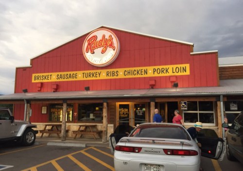 Exterior of Rudy's BBQ restaurant with a red facade and a sign listing menu items like brisket, sausage, and chicken.