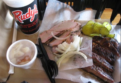 A plate of sliced brisket and ribs with pickles and onions, alongside a drink from Rudy's.