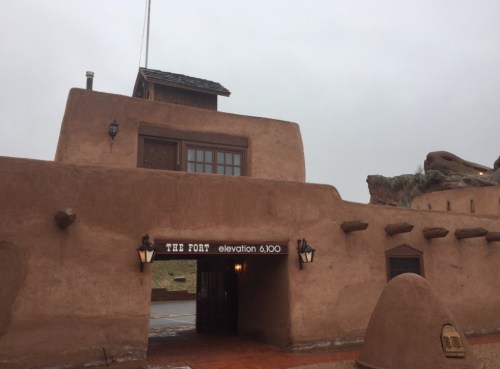 A rustic adobe building with a sign reading "The Fort" and an elevation marker, set against a cloudy sky.