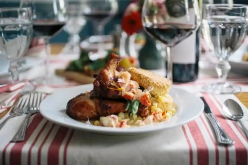 A plate of chicken with a side of salad and cornbread, set on a table with wine glasses and floral decor.