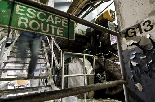 A person descends a metal staircase marked "Escape Route" near industrial machinery and a damaged wall.