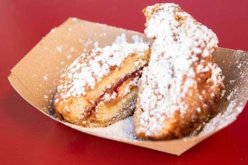 A close-up of a powdered sugar-dusted pastry filled with jam, served in a small cardboard tray.