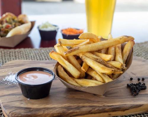 A serving of golden French fries in a paper bowl, accompanied by a small cup of dipping sauce on a wooden platter.