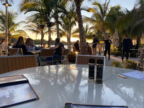 Outdoor dining scene with palm trees, sunset, and people enjoying a meal at a restaurant. Tables and menus visible.