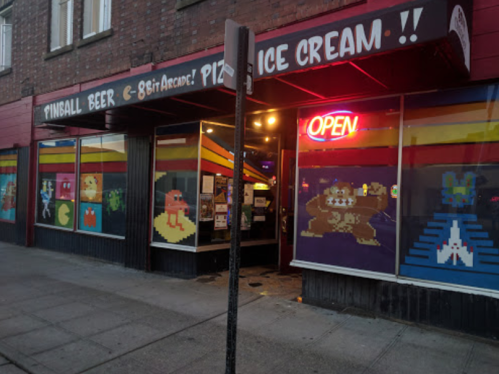 A colorful arcade and pizzeria with neon "OPEN" sign and retro video game artwork on the windows.