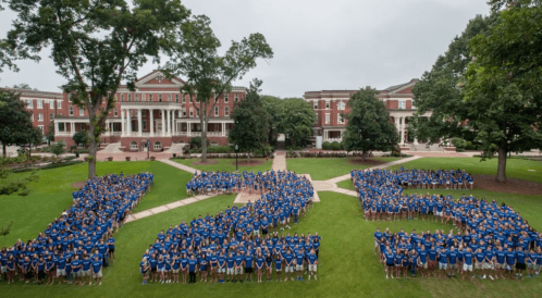 A large group of students in blue shirts forms the number "125" on a grassy campus area, surrounded by buildings and trees.