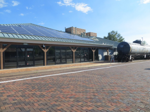 A building with solar panels beside a train track, featuring a tank car and a brick pathway in front.