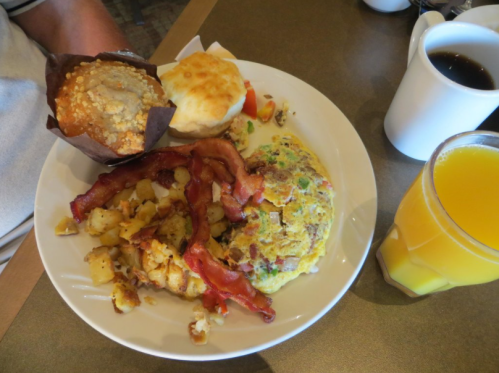 A plate of breakfast featuring an omelet, bacon, potatoes, a biscuit, a muffin, and a glass of orange juice.