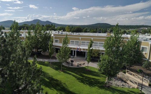 Aerial view of a building surrounded by trees and grass, with mountains in the background under a partly cloudy sky.