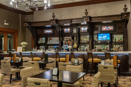 A stylish bar area with a wooden counter, high stools, and a variety of drinks displayed on shelves.