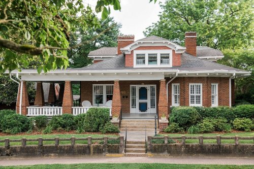 A charming brick house with a front porch, surrounded by greenery and trees, featuring a blue door and white trim.