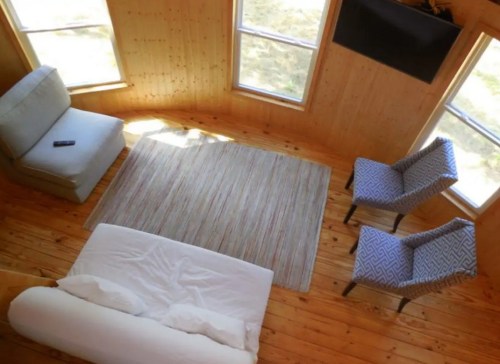 Cozy wooden interior with a sofa, two patterned chairs, a rug, and large windows letting in natural light.