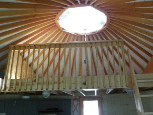 Interior view of a yurt with a circular skylight and wooden beams, featuring a balcony railing above.