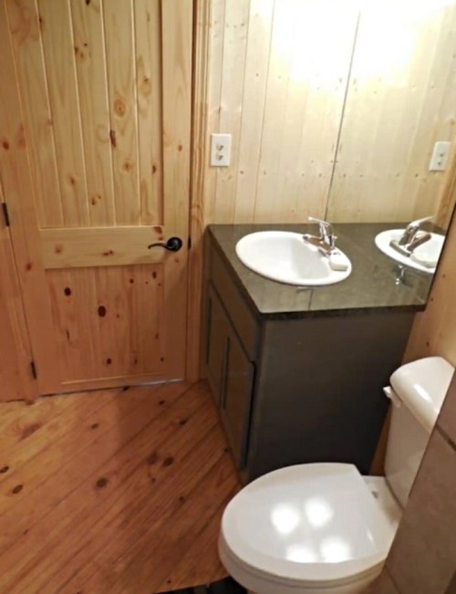 A small bathroom featuring a wooden door, sink with a mirror, and a toilet on a wooden floor.