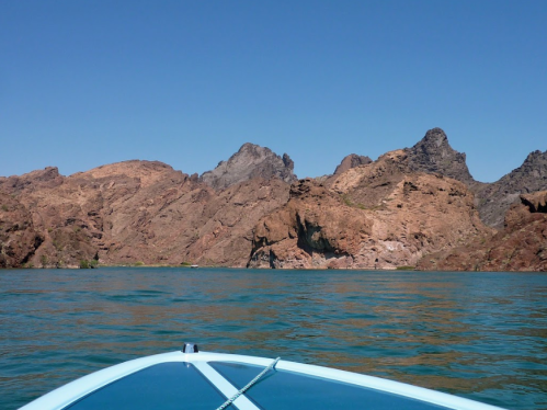 A view from a boat on calm water, showcasing rocky mountains under a clear blue sky.