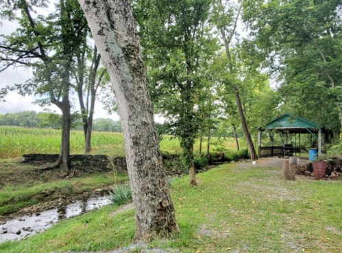 A serene outdoor scene featuring a tree-lined path, a gazebo, and a small stream beside a cornfield.