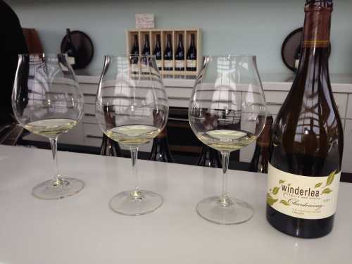 Three wine glasses filled with white wine, alongside a bottle labeled "Winderlea Chardonnay" on a bar counter.