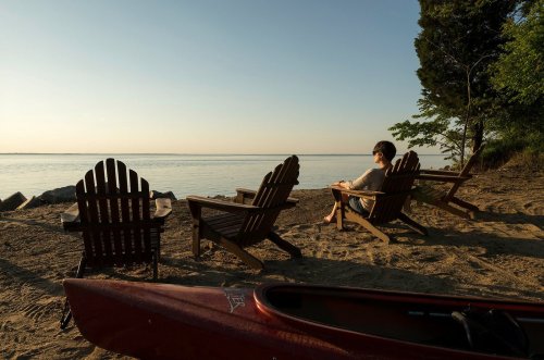 A person sits on a beach chair by the water, with kayaks and trees in the background under a clear sky.