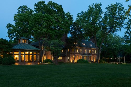 A large brick house with illuminated windows, surrounded by trees, at dusk with a well-maintained lawn.