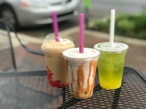 Three colorful drinks in clear cups with straws sit on a black table, with a blurred background of a street.