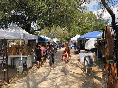 A bustling outdoor market with vendors under tents, people browsing art and crafts along a tree-lined path.