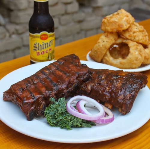 A plate of grilled ribs with onion slices and kale, accompanied by a bottle of Shiner Bock and fried onion rings.